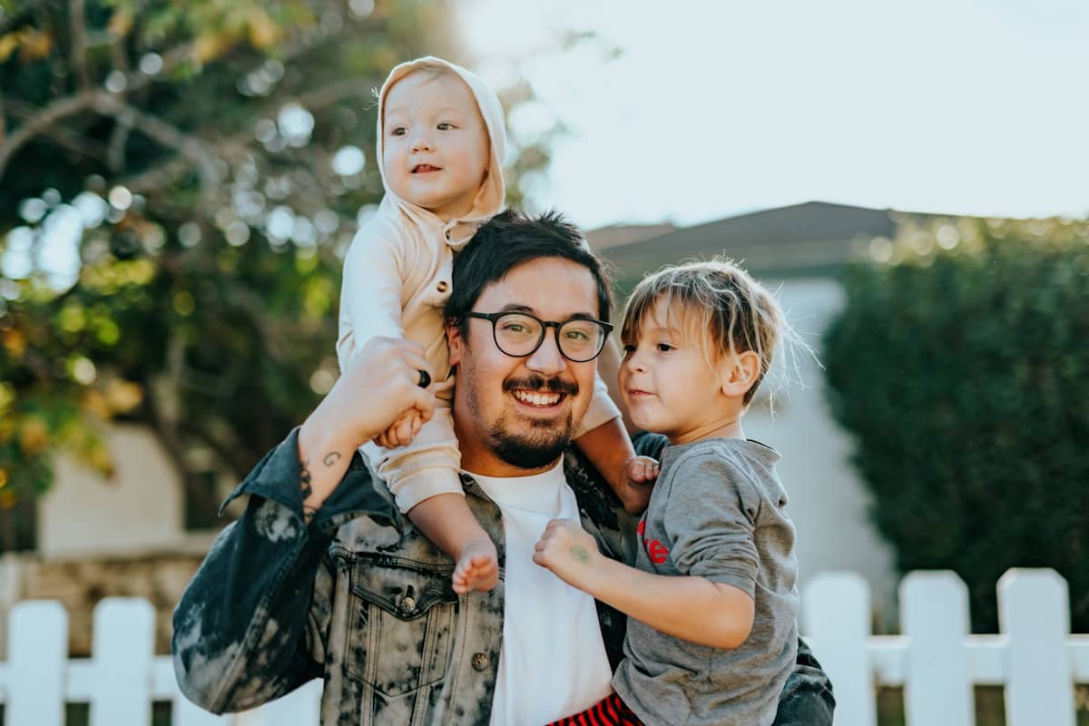 A dad with two young kids in a sunny front yard with a white picket fence
