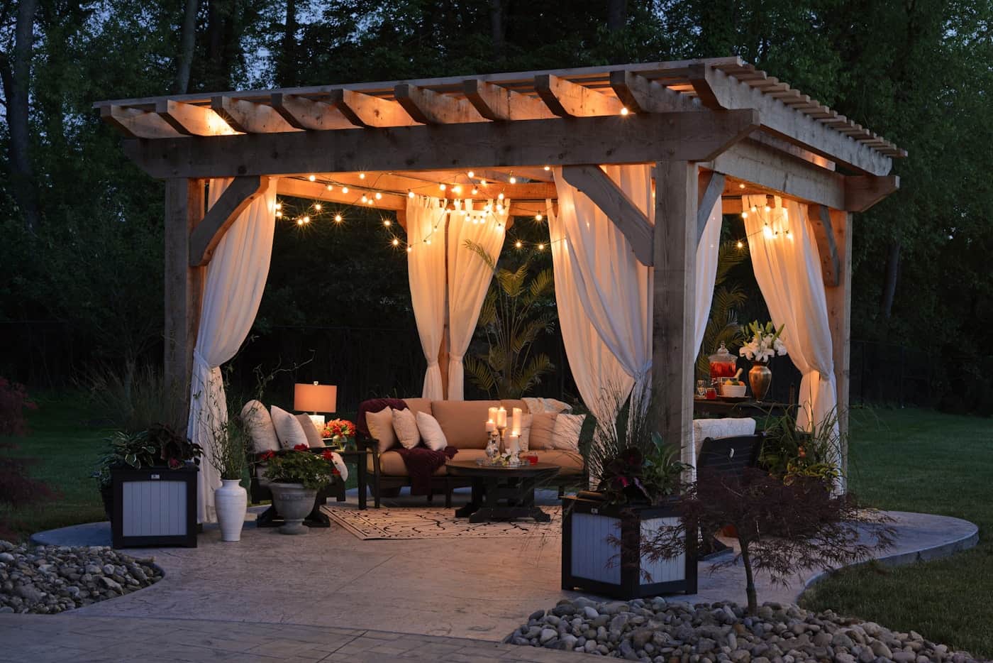 Backyard pergola strung with lights glowing at dusk over a cushioned family seating area